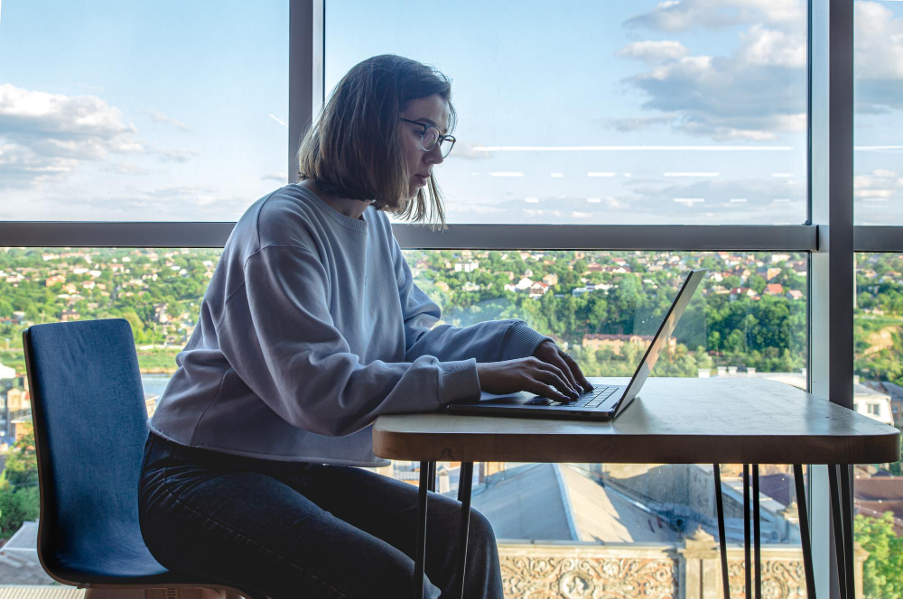 mulher jovem, de cabelos curtos, usando óculos, trabalha usando notebook. ao fundo há uma grande janela com vista para uma área verde, de natureza.