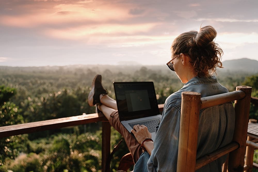 mulher jovem trabalha com o laptop sobre o colo, os pés sobre um guarda corpo. o ambiente é rústico e em volta há muita natureza.